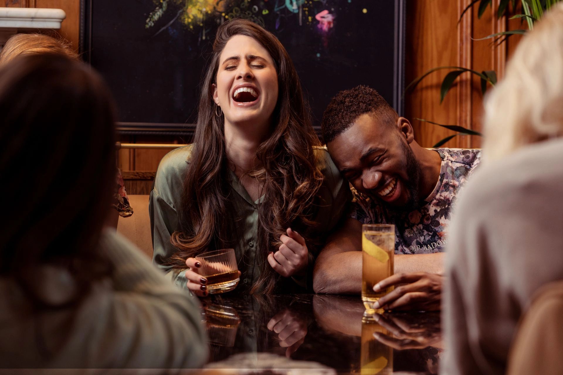 Two friends laugh heartily over drinks at a cozy gathering.