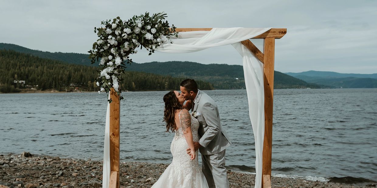 Bride and groom kissing under floral arch by the lake.