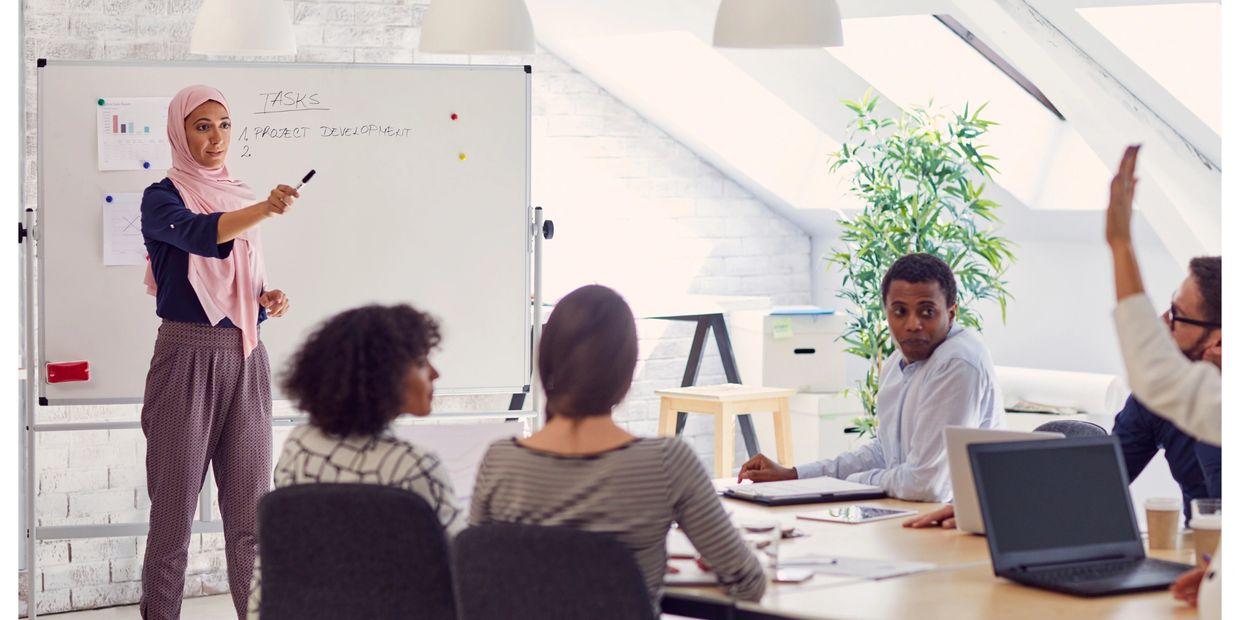 Photo of a diverse group of people at work with a woman near the whiteboard pointing to someone who 