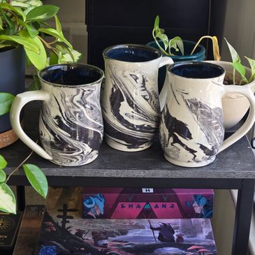 Three black and white marbled ceramic mugs on a shelf surrounded by plants.