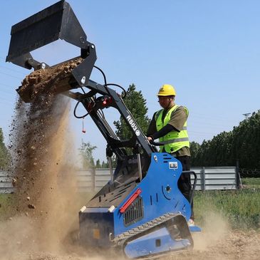 Worker operating a mini loader dumping soil outdoors on a sunny day.