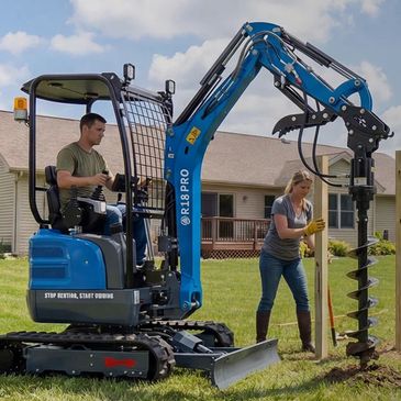 A man operates a mini excavator while a woman installs fence posts in a suburban yard.