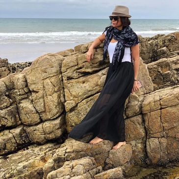 Woman in hat and sunglasses standing barefoot on rocky beach.