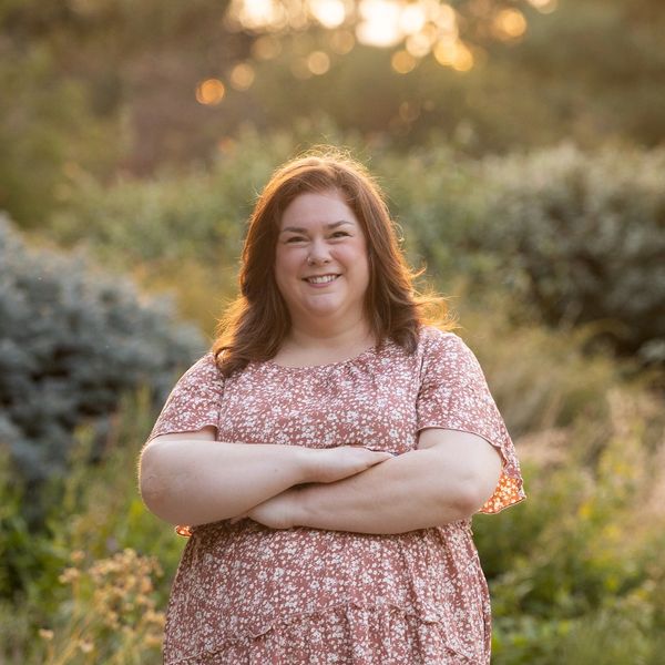 Smiling woman in a floral dress stands outdoors with arms crossed in a sunlit natural setting.