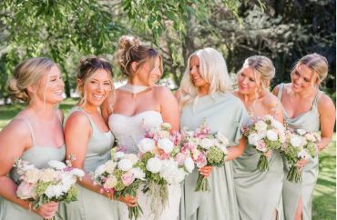 Bridesmaids in sage green dresses with bouquets, smiling outdoors.