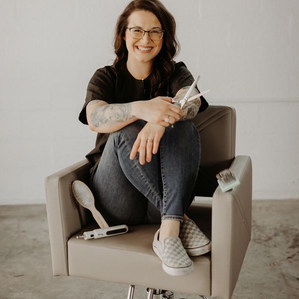 Smiling woman with glasses sitting on a salon chair holding scissors.