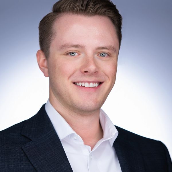 Young professional man in a dark suit smiling against a light background.
