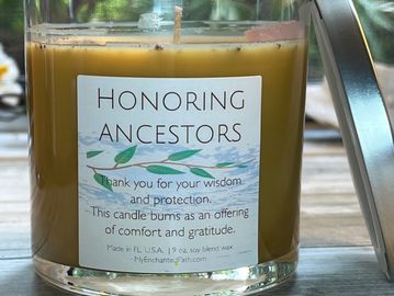 Glass holder filled with tan wax. The square label says honoring ancestors. there is a silver lid. 