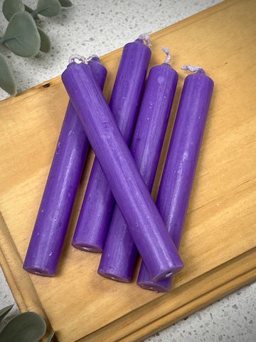 Five purple taper candles on a wooden tray with green leaves nearby.