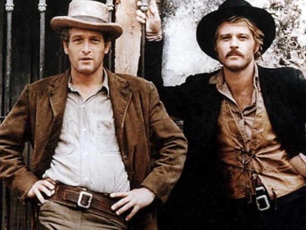 Two men dressed in vintage western attire leaning against a wall.