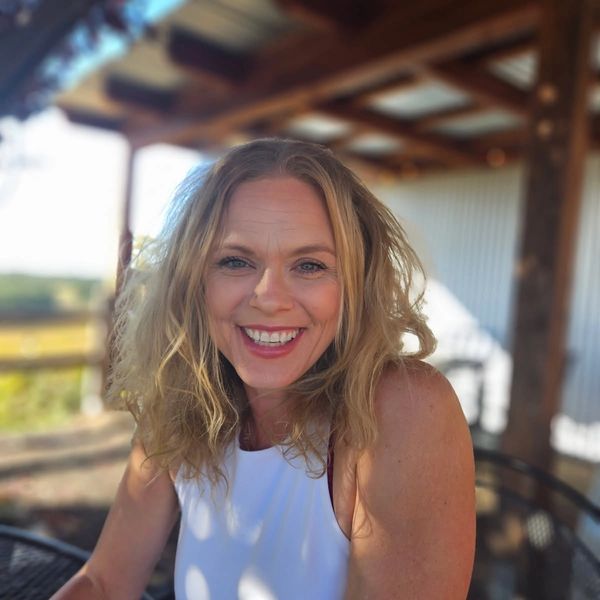 Smiling woman with curly hair in a sunlit outdoor setting.