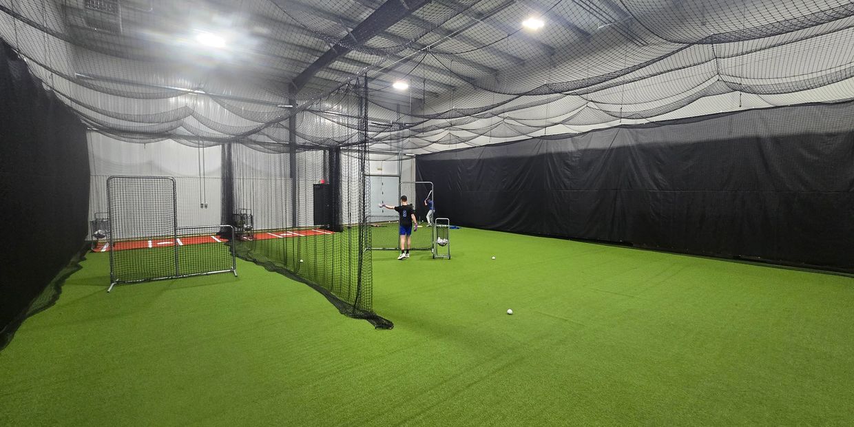 Indoor baseball training facility with nets and two people practicing.