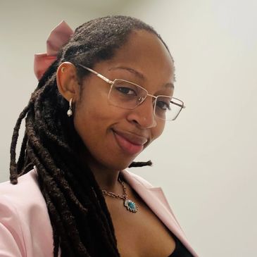 A woman with glasses and long dreadlocks smiles gently at the camera.