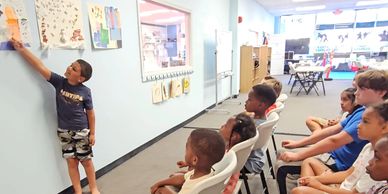 A young boy presents to a seated classroom of attentive children.