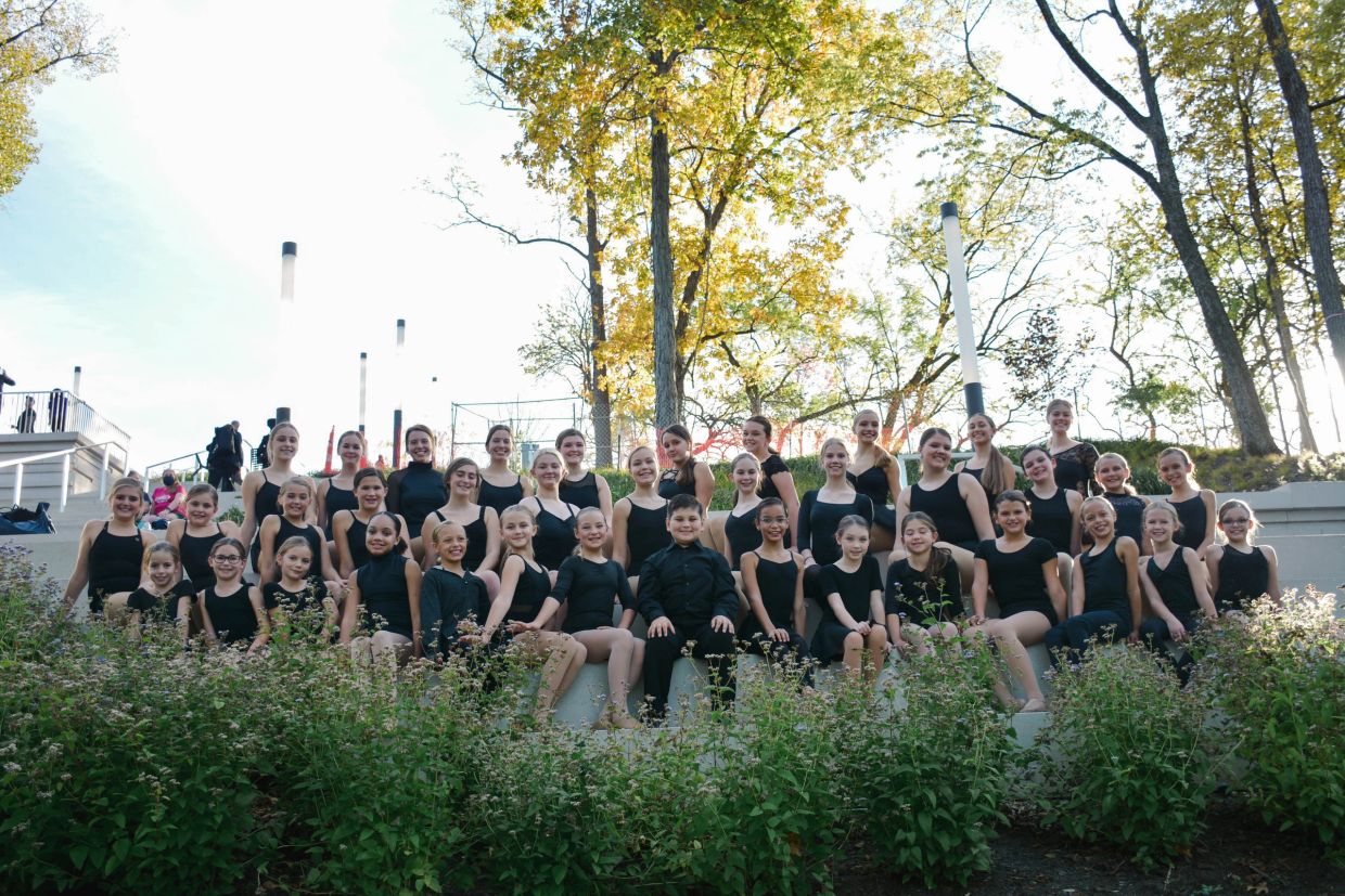 Large group of dancers in black attire posing outdoors on stone steps.