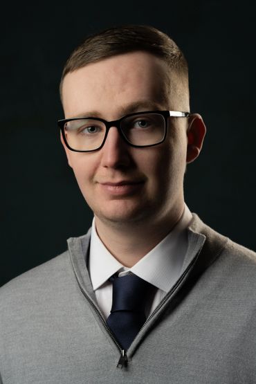 Portrait of a man wearing glasses, a gray sweater, white shirt, and navy tie.