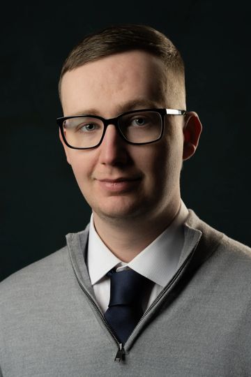 Young man with glasses wearing a grey sweater and navy tie.