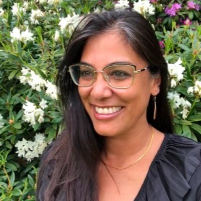 Smiling woman with glasses in front of flowering bushes.