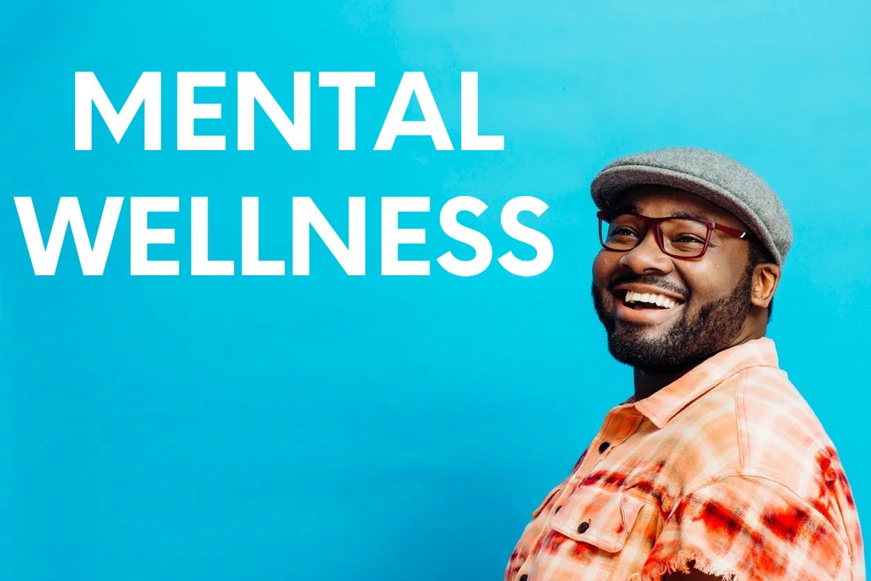 Black man smiling; wearing glasses and hat, orange & red pattern shirt, blue background wall.