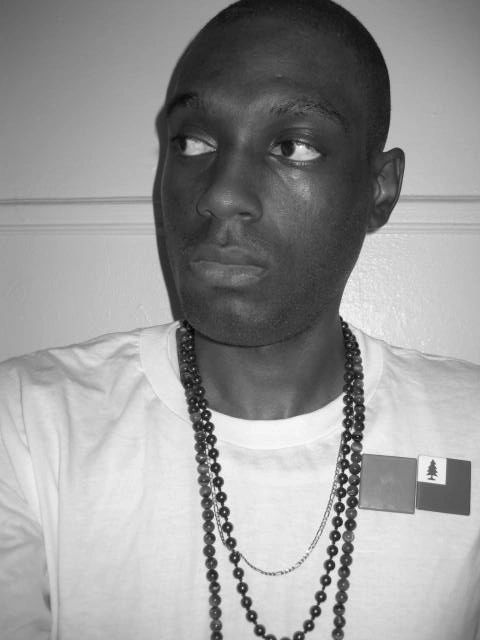 Black and white picture of Moorish Brooklyn, white t-shirt, mala beads, chain and two flag pins.