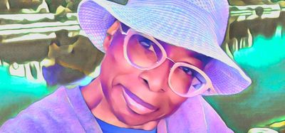 A woman wearing a hat and glasses smiles near a scenic water body.