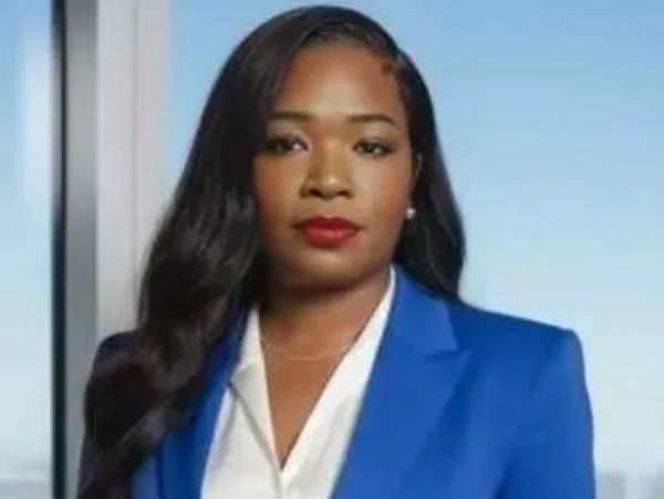 Confident woman in a blue blazer and white blouse poses indoors with a city view.