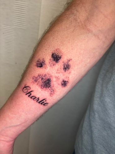 Tattoo of a black paw print with the name Charlie underneath on a forearm.