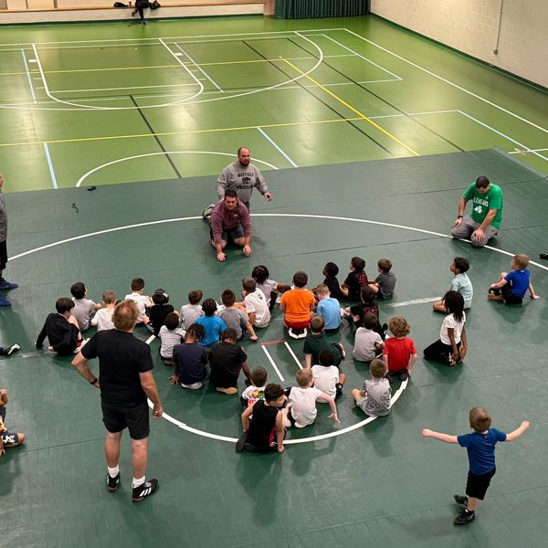 Kids gathered on wrestling mats with coaches in gym.