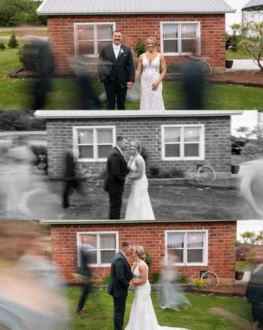 A bride and groom pose in front of a brick house with blurred wedding guests around.