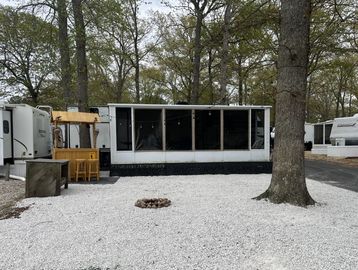A white mobile home with a screened porch and gravel yard in a wooded area.