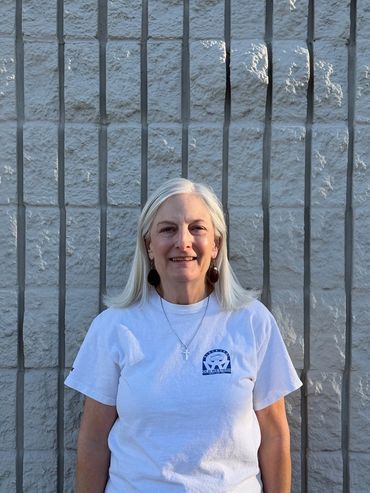 Smiling woman with long gray hair in a white t-shirt standing against a gray textured wall.