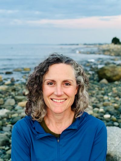 Smiling woman with curly hair by rocky shoreline.