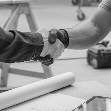 Two people wearing gloves shaking hands over a work table with blueprints and a tablet.
