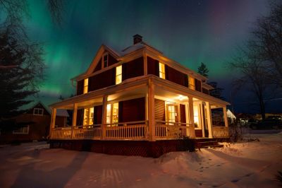 A warmly lit house under a vibrant aurora borealis in a snowy landscape at night.