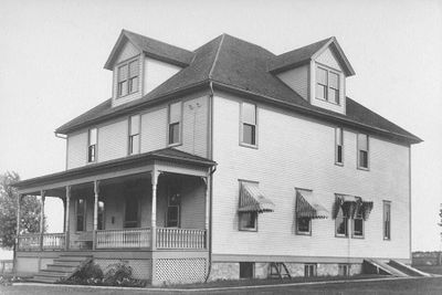 Black and white photo of a large two-story house with a porch and dormer windows.