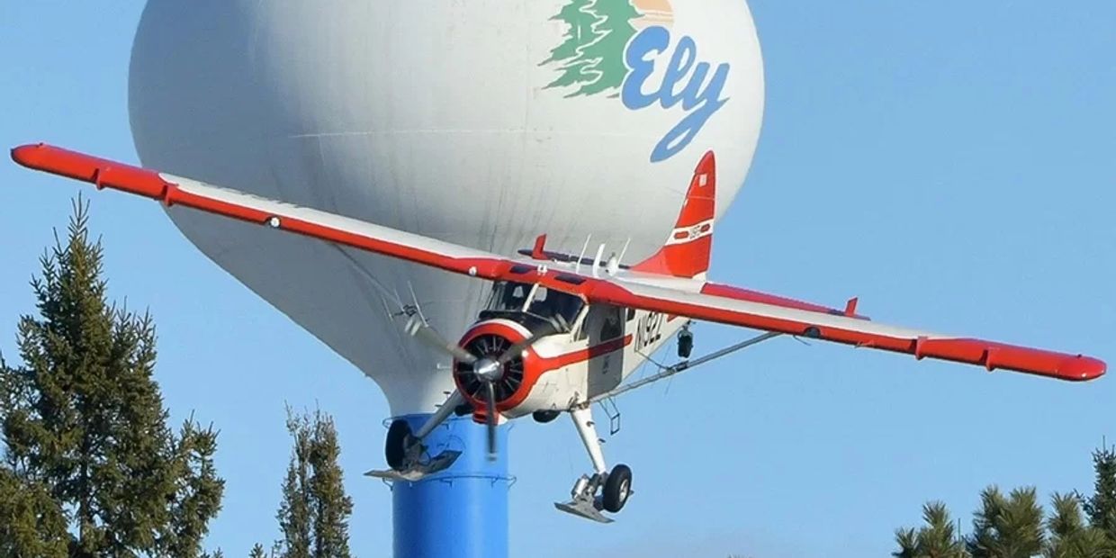 A small red and white plane flies near a water tower with the word Ely.