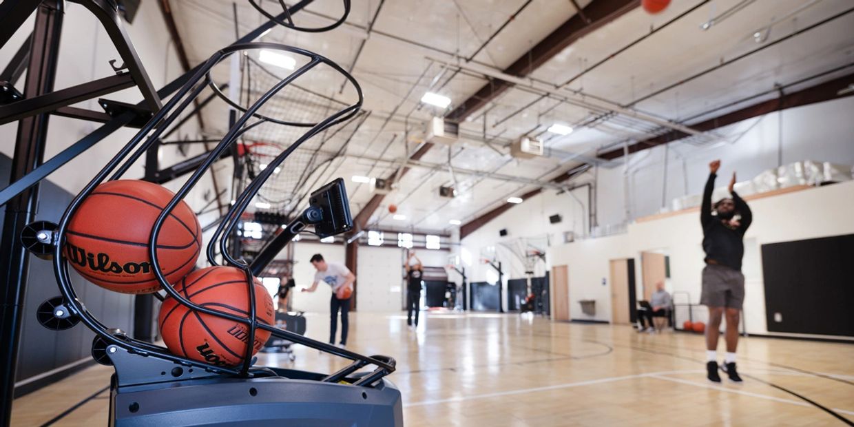 People practicing basketball shooting with a ball return machine in an indoor court.