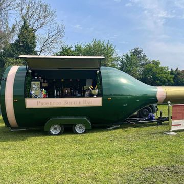 A large green prosecco bottle-shaped bar trailer outdoors on grass.