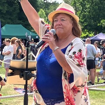 Woman in floral kimono and hat singing into microphone at outdoor event.