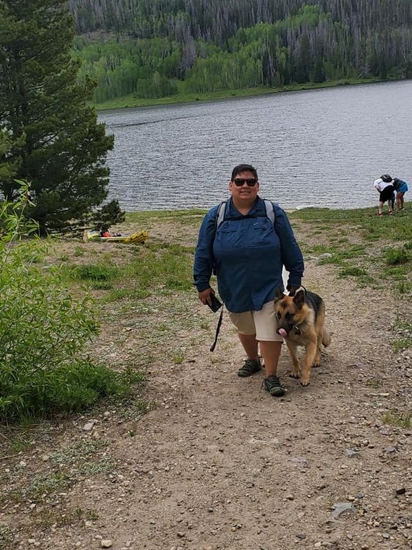 Person walking a German Shepherd dog near a lake in a forested area.
