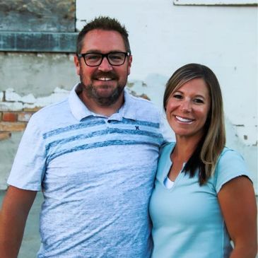Smiling couple standing close together outdoors against a weathered wall.