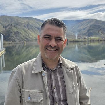Smiling man standing by a serene lake with mountains in the background.