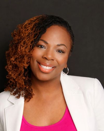 Smiling woman with curly hair wearing white blazer and pink top.