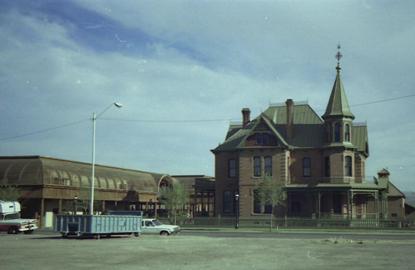 Historic brick house with pointed tower next to a modern building and cars.