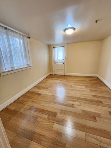 Empty room with wooden floor, beige walls, window with curtains, and a door.