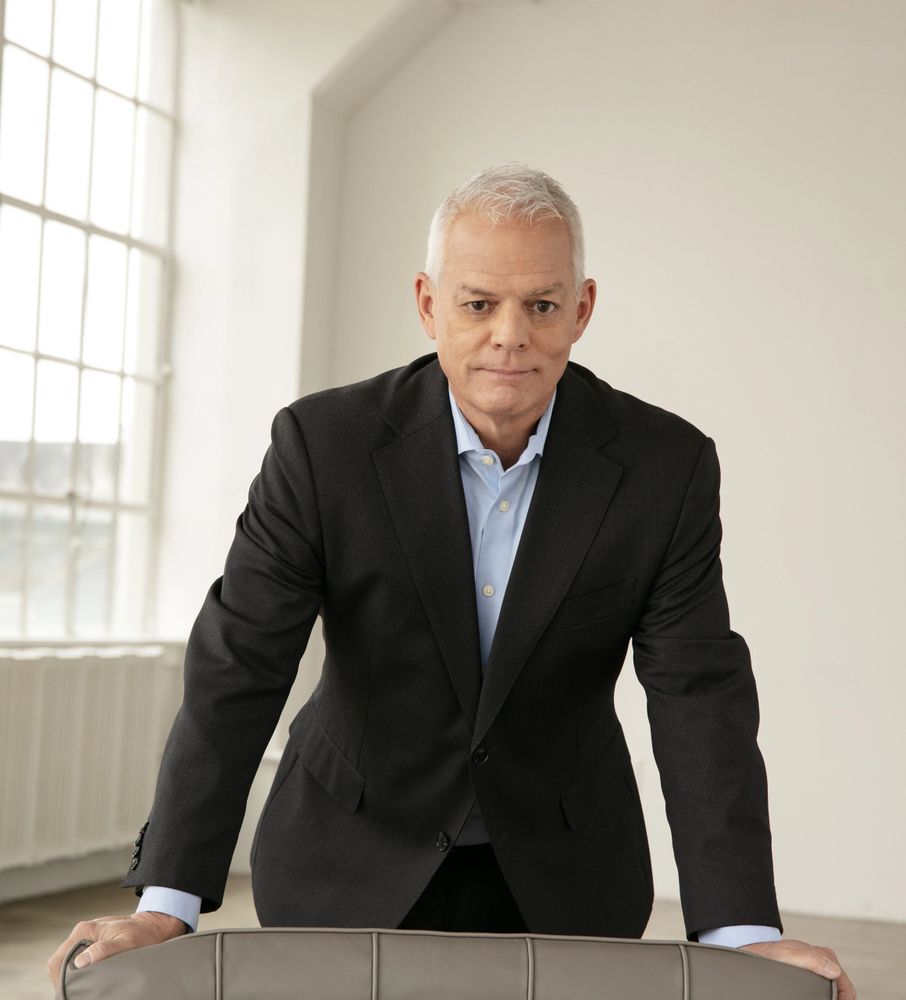 Confident middle-aged man in a suit leaning on a chair in a bright room.