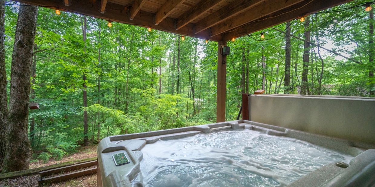 Outdoor hot tub under wooden roof with forest view and string lights.