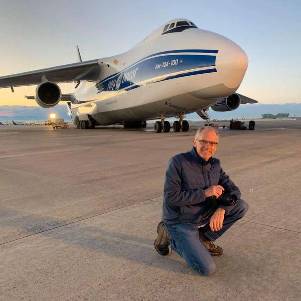 Ken kneeling with his camera in his hand on a tarmac in front of an Antonov An- cargo transport airp