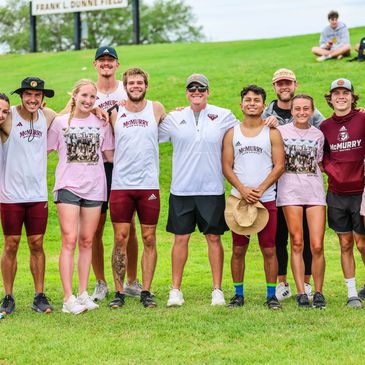 A group of smiling McMurry University track and field athletes and coach posing outdoors on a grassy field.