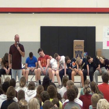 A man speaking as several people appear asleep on chairs in front of an audience.